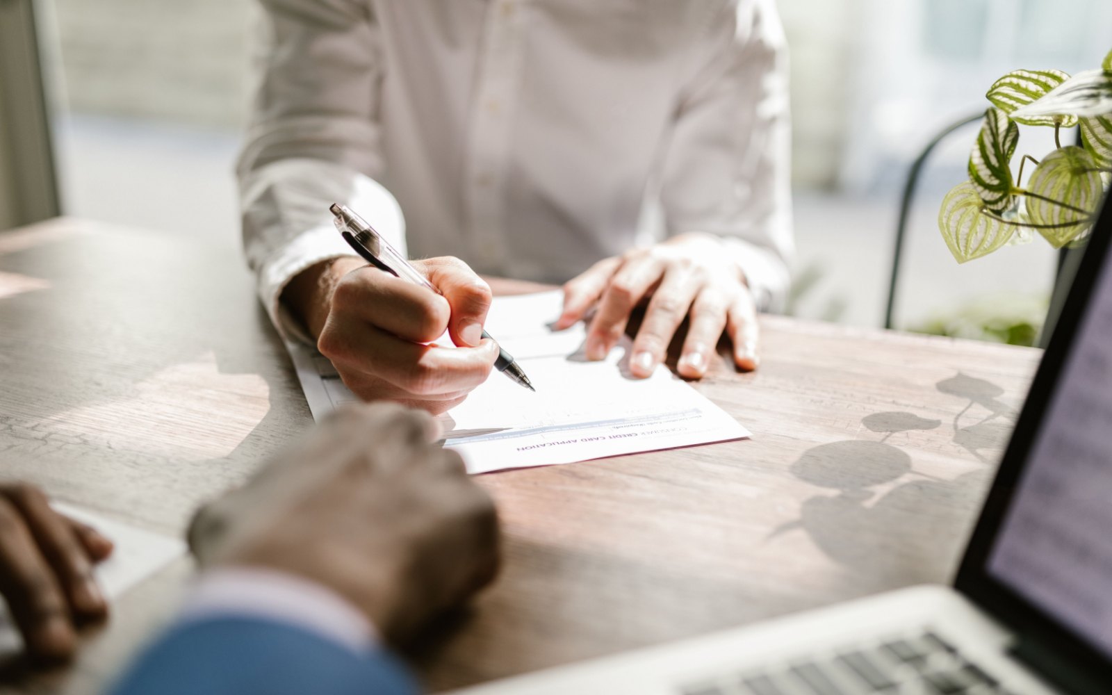 Two people at a wooden table, one filling in a document with a pen while the other looks on, a laptop at the edge