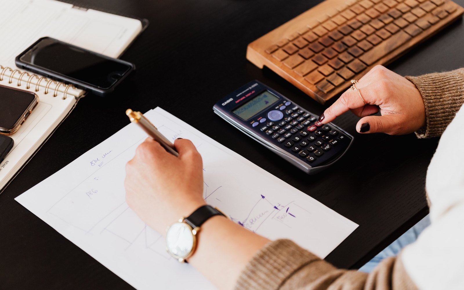 Hands writing figures on a sheet of paper and typing on a calculator at a desk with a notebook and a phone